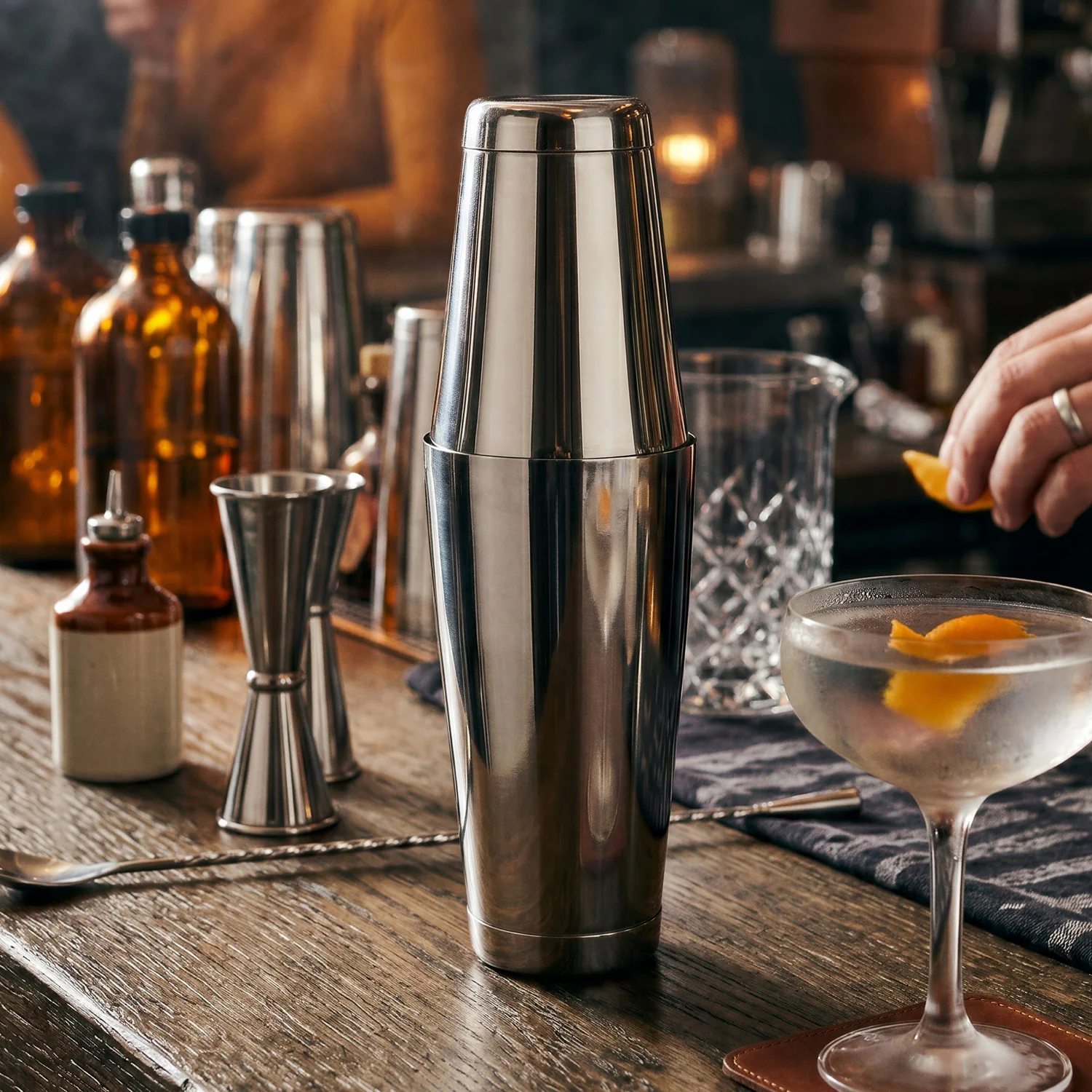 A stainless steel cocktail shaker sits on a wooden bar alongside a jigger, glass mixing jug and a chilled cocktail garnished with orange peel. A stainless steel cocktail shaker sits on a wooden bar alongside a jigger, glass mixing jug and a chilled cocktail garnished with orange peel.